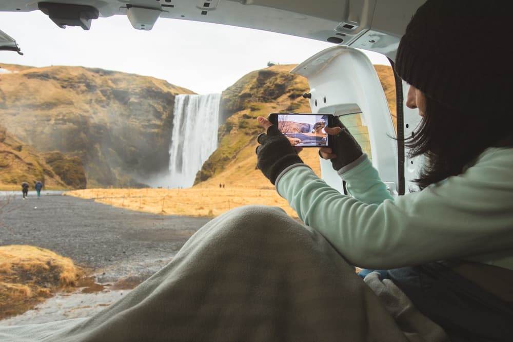 Women sitting in a camper van taking a photo of Skogafoss waterfall.