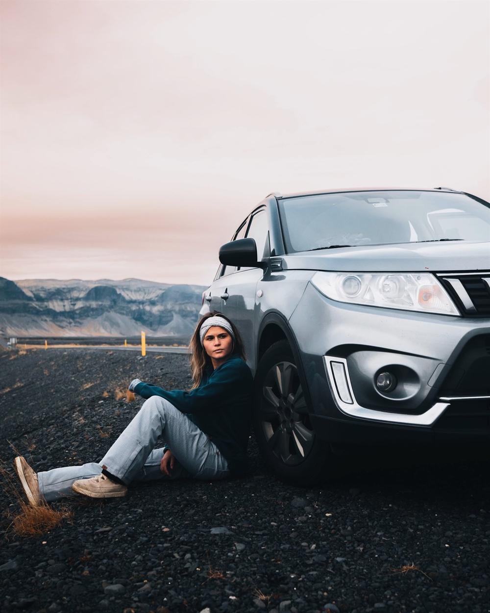 Femme assise près de son 4x4 Suzuki Vitara loué chez Blue Car Rental, dans la campagne islandaise.