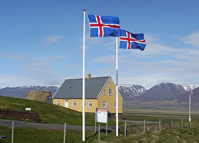 Icelandic flags flying beside a yellow farmhouse with snow capped mountains behind