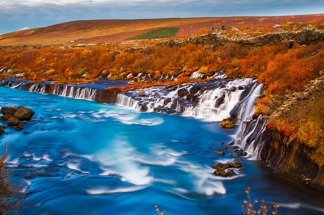 Autumn colours along an Icelandic waterfall. Waterfall cascading into a blue river surrounded by red autumn shrubs in Iceland