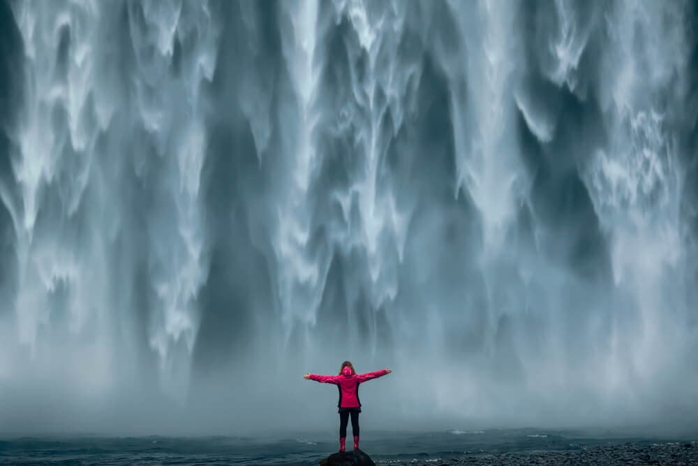Iceland top reasons to visit waterfall. Person standing in front of a beautiful waterfall in Iceland.